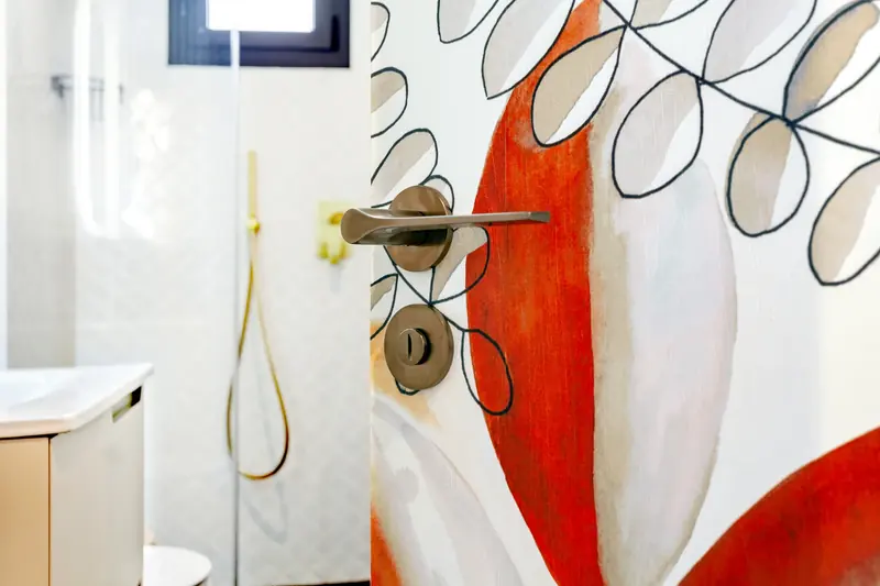Detalle de baño con grifería dorada y mural de hojas en tonos rojos